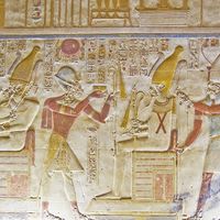 An ancient egyptian hieroglyphic painted carving showing the falcon headed god Horus seated on a throne and holding a golden fly whisk. Before him are the Pharoah Seti and the goddess Isis. Interior wall of the temple to Osiris at Abydos, Egypt.